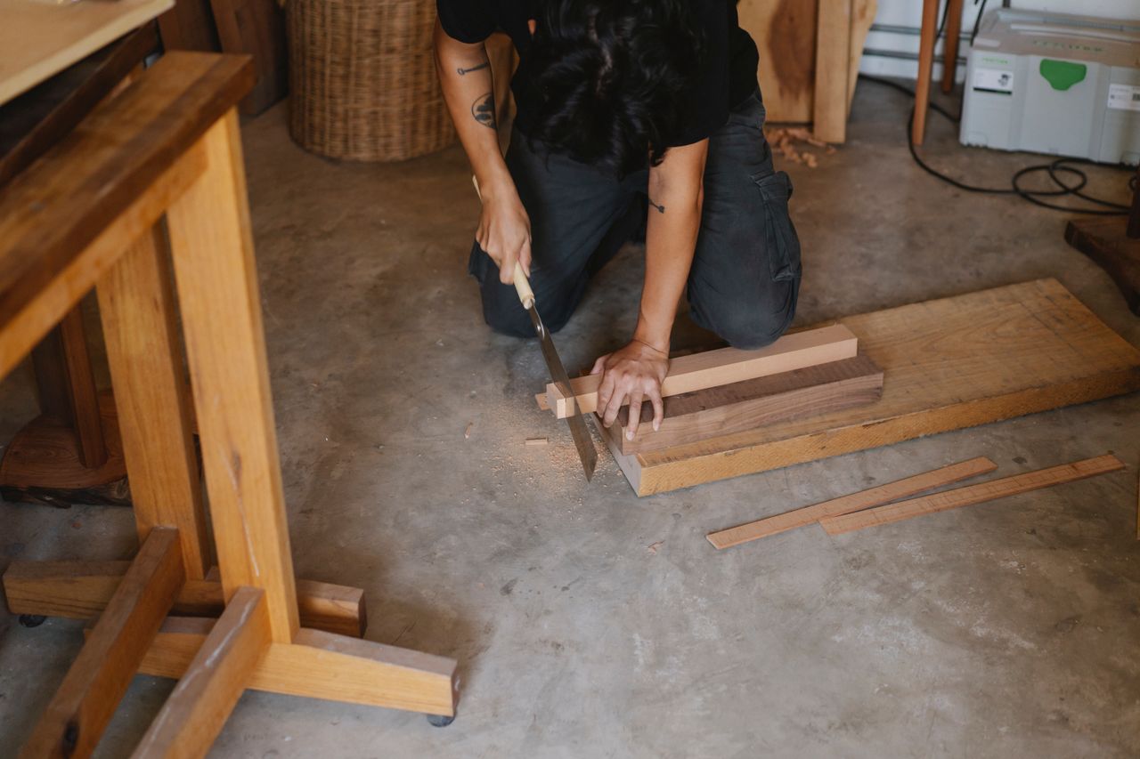 Anonymous joiner working with Japanese saw while cutting edge of timber plank on floor in workshop